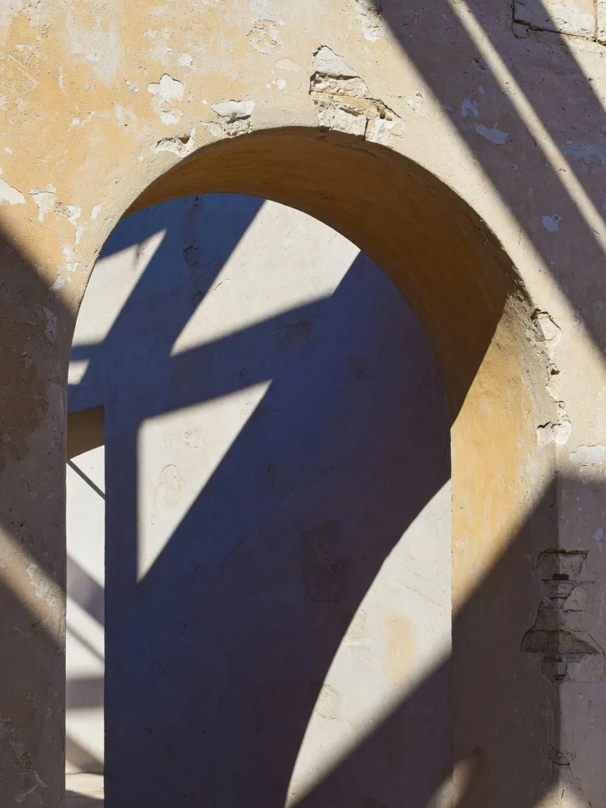 A weathered stucco archway in harsh midday sun, casting razor-sharp geometric shadows across its curved recess