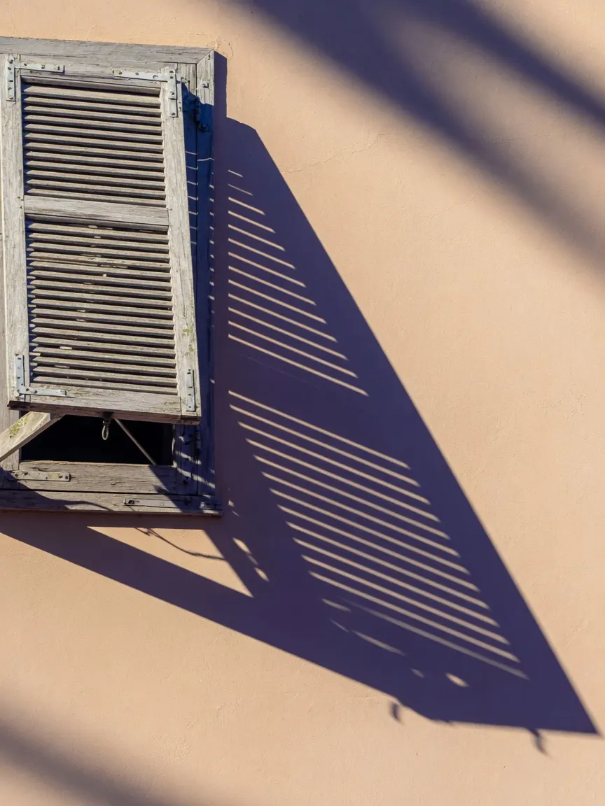 A half-open louvered shutter of bleached cypress, its angled slats carving hard diagonal blades of subtropical shadow across a smooth stucco wall, empty and severe