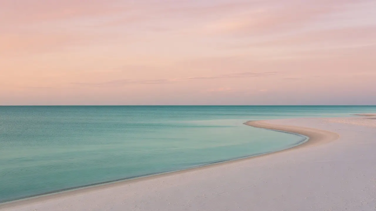 Wide Gulf coast vista at dawn: empty sugar-sand beach curving into glassy turquoise water beneath a vast, hazy rose sky, horizon razor-sharp, no people or objects