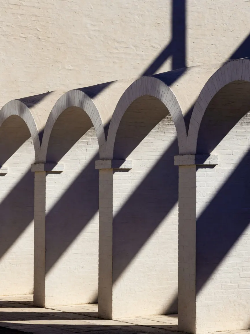 A crisp colonnade of whitewashed brick arches casting razor-sharp diagonal shadows across a sun-bleached stucco wall, empty and severe